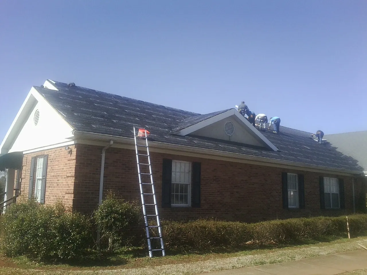 Crew tearing shingles off a brick ranch roof with a ladder set — Emergency Roof Repair work by our crew in Owens Cross Roads
